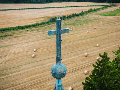 Aerial view of Arlava Evangelical Lutheran Church spire with a copper cross and orb set against a vast harvested field with hay bales in Valdemarpils, Talsi Municipality, Latvia.