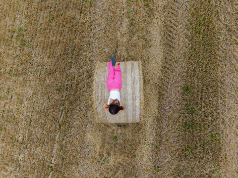 Aerial view of a woman in pink pants and a white top lying on a round hay bale in a harvested field in Latvia.