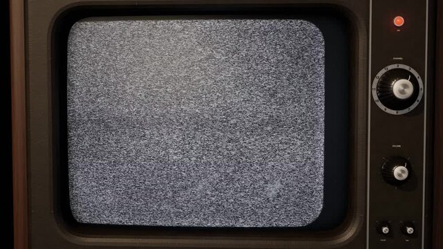 Vintage CRT television screen filled with analog static noise, wood cabinet and metal tuning dials in a dark room, seamless looping retro TV background