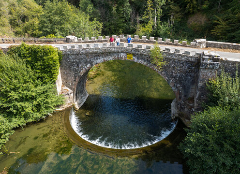 Aerial view of the historic stone bridge over the River Teme, with its circular weir, surrounded by lush green trees, with people standing on the bridge in Ludlow, Shropshire, United Kingdom.