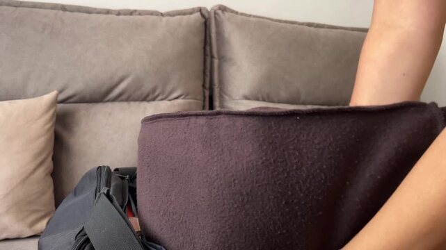 Close-up of hands placing folded fleece apparel into a travel bag on a couch. Suitable for luggage commercials, travel prep content, gear packing, and organization guides.