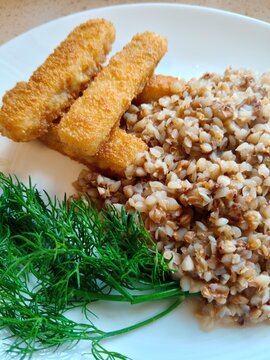 Homemade Buckwheat Groats Served with Breaded Fish Sticks and Dill. Close-up of cooked buckwheat groats served alongside several breaded fish sticks and fresh dill on a white plate. The dish shows 