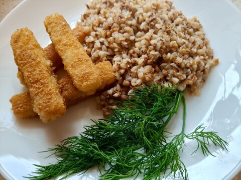 Simple Homemade Meal of Buckwheat, Breaded Fish Sticks and Dill. Homemade meal featuring cooked buckwheat groats, three breaded fish sticks and fresh dill served together on a white plate. The composi