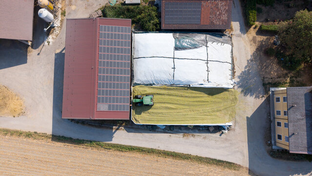 Aerial view of a tractor working on a large silage storage pit at a livestock farm, featuring covered forage and solar panels on the barn roof in Milan, Lombardy, Italy.