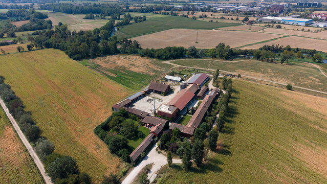 Aerial view of a traditional Italian farm with terracotta-roofed buildings, a central courtyard, and rooftop solar panels, set amidst expansive agricultural fields in Mediglia, Lombardy, Italy.