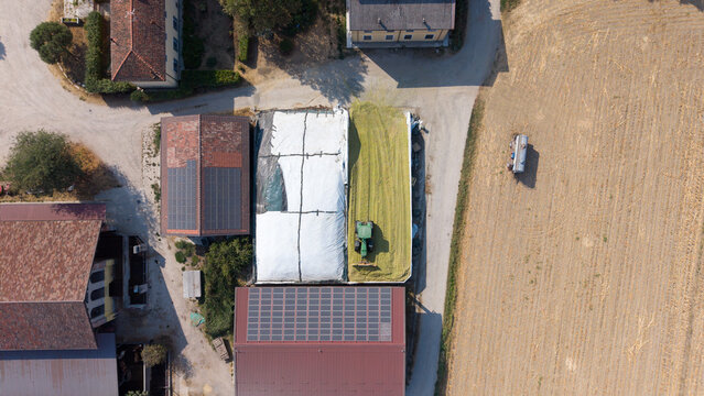 Aerial view of a tractor working on a silage storage pit at a livestock farm with solar panels on nearby barn roofs in Milan, Lombardy, Italy.