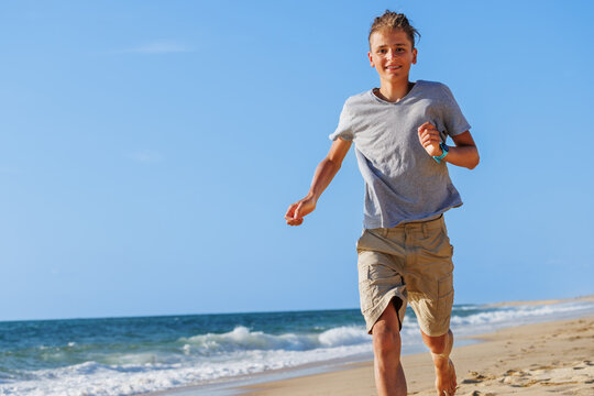Teenage boy jogging barefoot along a sunny beach beside ocean