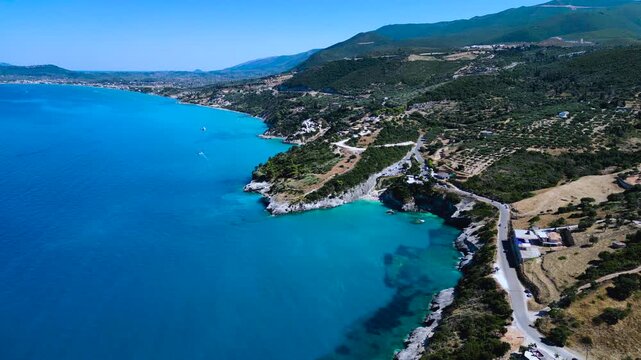 Drone flies over rugged Mediterranean coastline with crystal turquoise water, winding coastal road, and hillside village.