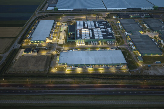 Aerial view of a large hyperscale AI data center complex illuminated at night, featuring industrial buildings, cooling infrastructure, and surrounding fields in the Netherlands.