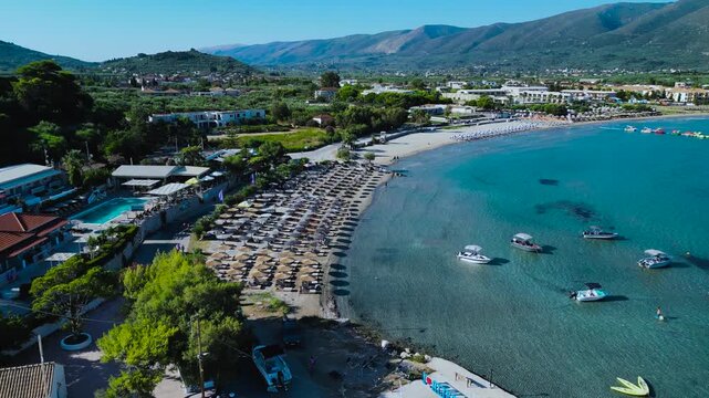Aerial zoom out reveals packed beach with straw umbrellas, crystal turquoise water, boats, and resort hotels backed by green hills.