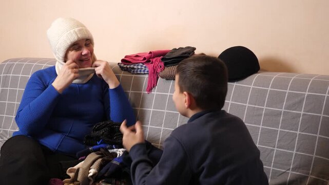 Mother tries on a warm winter hat and scarf, adjusts them and shows her son the clothing during household activities.