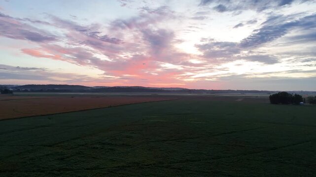 A drone captures a wide freshly plowed farm field bordered by a green pasture and a quiet rural road as vivid pink sunset clouds stretch across the sky above distant rolling hills.