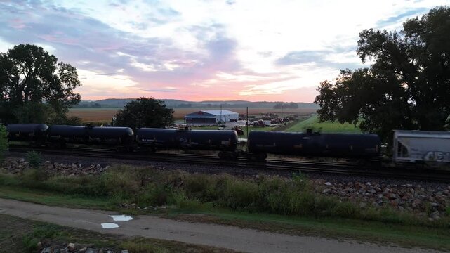 A drone tracks alongside a long freight train hauling tanker and hopper cars as it rolls through open farmland beneath a fading pink sunset sky near a quiet country road.