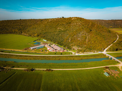 Aerial view of a farm complex situated along a river with surrounding agricultural fields and a forested hill in the background in Brtonigla, Istria County, Croatia.