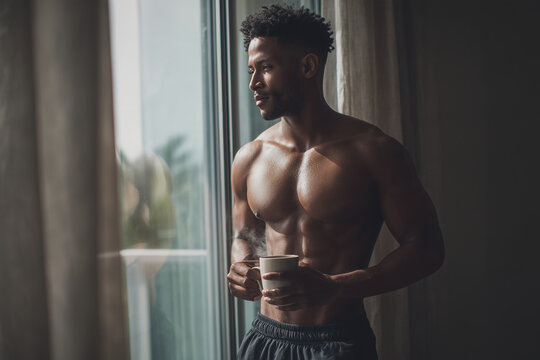 Shirtless African American Man Holding Coffee Mug By Large Window In Morning Light At Home