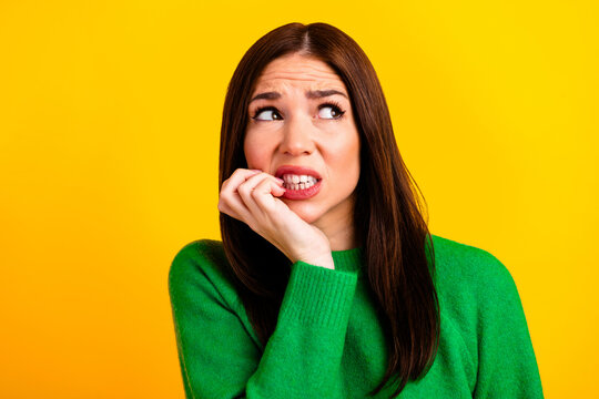 Nervous young woman biting her nails and looking worried and anxious while wearing a green sweater against a bright yellow background