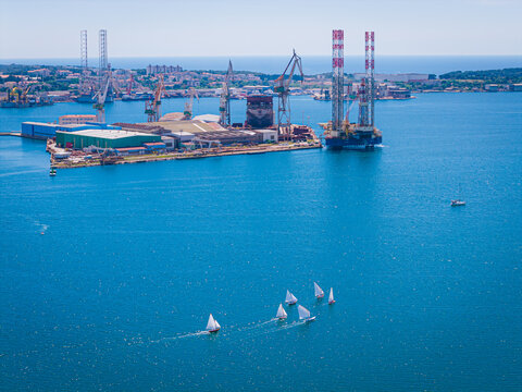 Aerial view of the Uljanik shipyard with industrial cranes and a jack-up rig docked in the harbor, accompanied by small sailboats on the blue Adriatic Sea in Pula, Istria County, Croatia.