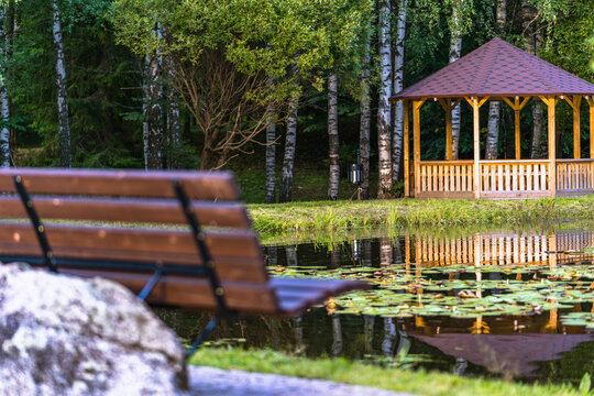 View of a wooden gazebo by a pond with water lilies and a wooden bench in the foreground surrounded by birch trees in Latvia.