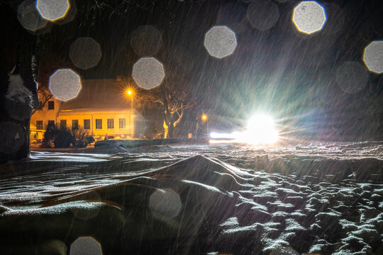 View of a snow-covered street at night with heavy snowfall, bright streetlights, and an illuminated building in the background in Latvia.