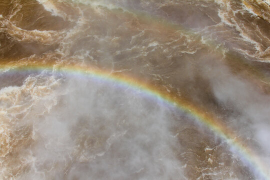 View of a vibrant rainbow arching over the powerful rushing water and mist of Iguazu Falls in Foz do Iguacu, Parana, Brazil.