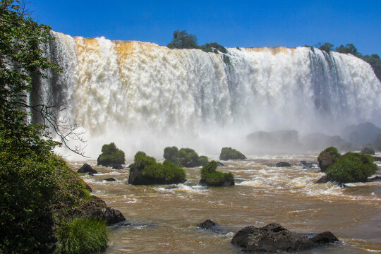 View of the powerful Iguazu Falls with cascading water, mossy rocks in the foreground, and lush greenery under a clear blue sky in Foz do Iguacu, Parana, Brazil.