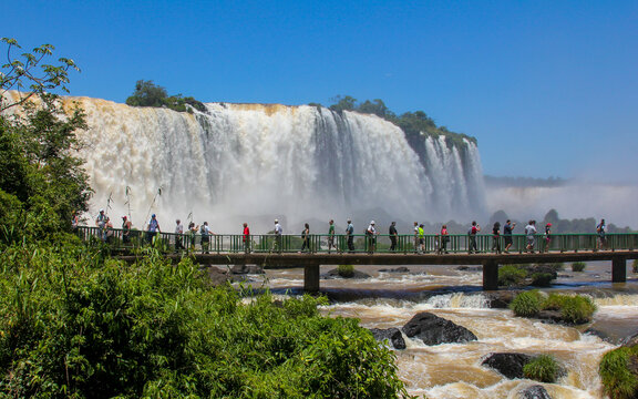 View of tourists walking on a metal walkway in front of the powerful Iguazu Falls surrounded by lush tropical vegetation under a clear blue sky in Foz do Iguacu, Parana, Brazil.