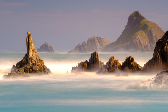 View of jagged sea stacks rising from the misty Atlantic ocean at low tide with golden light hitting the rock formations in Spain.