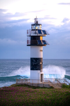 View of the black and white striped Faro de Isla Pancha lighthouse with solar panels, surrounded by purple flowers and crashing ocean waves in Spain.