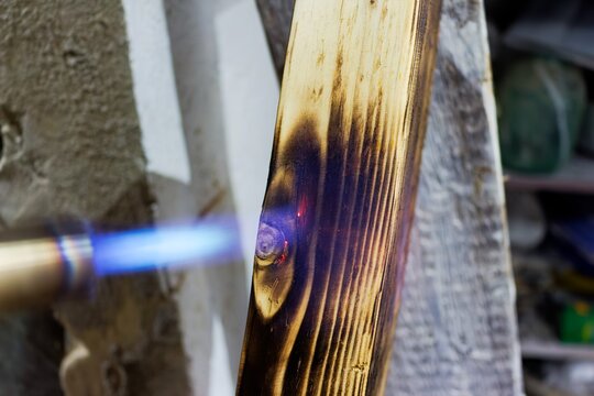 Close-up view of a craftsman using a gas torch to scorch and char a wooden board for decorative texture and wood preservation techniques in a workshop setting
