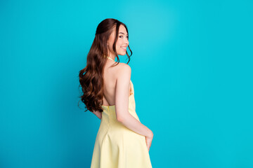 Smiling young woman in a yellow backless dress posing against a bright turquoise background, elegant profile portrait with long wavy hair and a joyful, confident expression © deagreez
