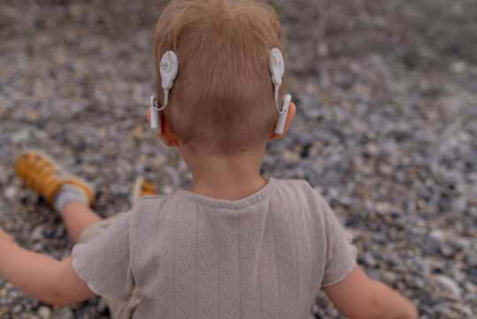 Rear view of a young child wearing bilateral cochlear implants while sitting outdoors on a pebble beach.