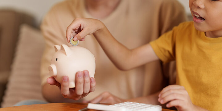 Asian boy putting coin in piggy bank with parent for financial literacy and savings habit