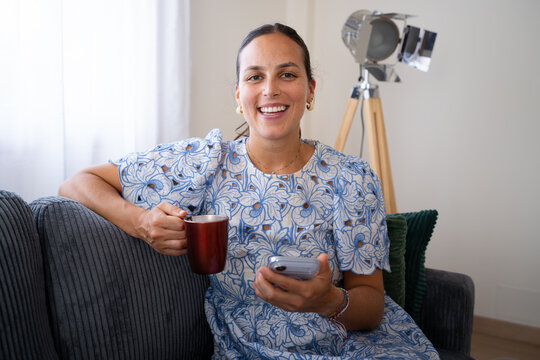 White woman smiling with mug and smartphone sitting on sofa indoors