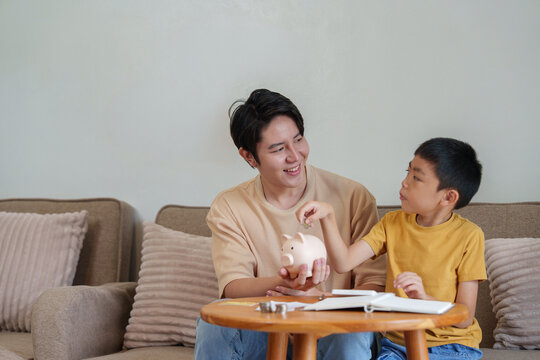 Asian father teaching young son how to save money using piggy bank at home