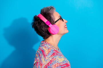 Smiling mature woman in pink headphones enjoying music against a bright blue background, colorful sweater and glasses in cheerful profile portrait © deagreez