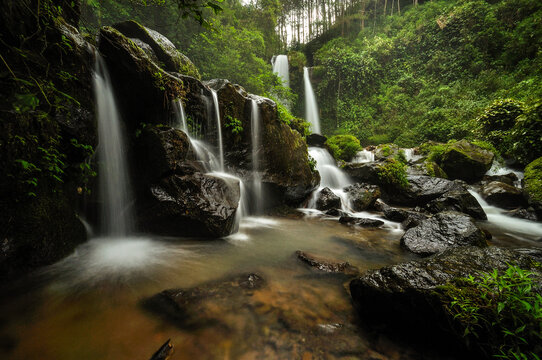 View of the cascading Air Terjun Grenjengan Kembar waterfall surrounded by lush green forest and dark wet rocks in Pakis, Central Java, Indonesia.