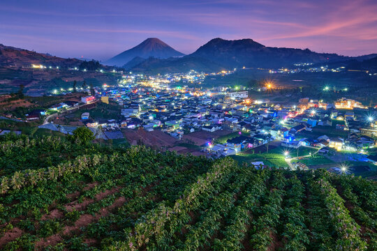 View of the illuminated town of Batur nestled in the Dieng Plateau with Bukit Skoter and mountains under a twilight sky, featuring agricultural fields in Batur, Central Java, Indonesia.