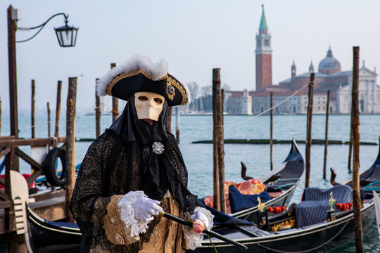 View of a person in an elaborate traditional Venetian carnival costume and mask standing by a gondola on a canal with San Giorgio Maggiore in the background in Venice, Veneto, Italy.