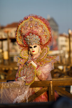 View of a person in an elaborate pink and gold Venetian carnival costume with an ornate mask and headpiece, holding a decorative fan behind a wooden railing in Venice, Veneto, Italy.