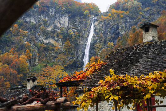 View of a tall waterfall cascading down a rocky cliff face behind traditional stone houses with slate roofs surrounded by vibrant autumn foliage in Switzerland.