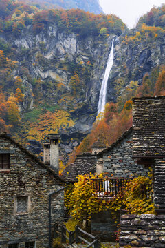 View of a tall waterfall cascading down a rocky cliff behind traditional stone houses with autumn foliage in Switzerland.