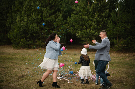 Ecstatic family celebrating twin gender reveal with flying confetti and balloons in a sunny forest clearing.