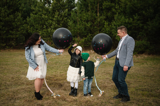 Happy family with two children holding black gender reveal balloons for twins outdoors in a pine forest.