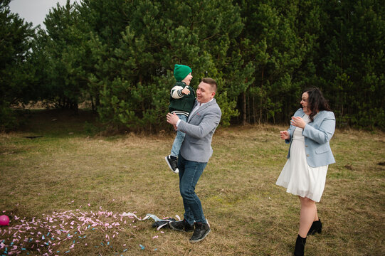 Happy father holding his laughing little son with a pregnant mother walking nearby on a nature clearing in a forest.