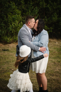 Loving parents kissing each other while their little daughter hugs them outdoors in a forest during a photo shoot.