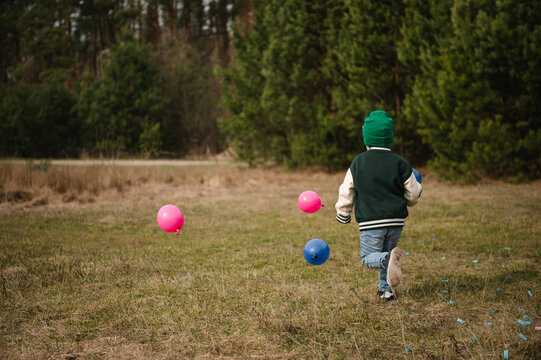 Little boy running with small blue and pink balloons across a clearing in a pine forest.