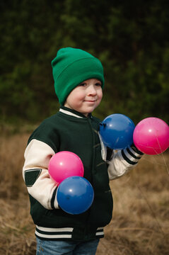 Cute little boy holding small blue and pink balloons outdoors in a forest during a family celebration.