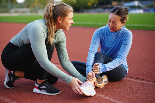 Injured runner sitting on a red track with a strained expression, holding her ankle while a concerned teammate crouches beside her, offering support and examining the injury