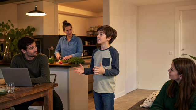 Family doing parallel activities at home while young boy speaks in living room. Parents and siblings in kitchen and working on laptop during evening routine