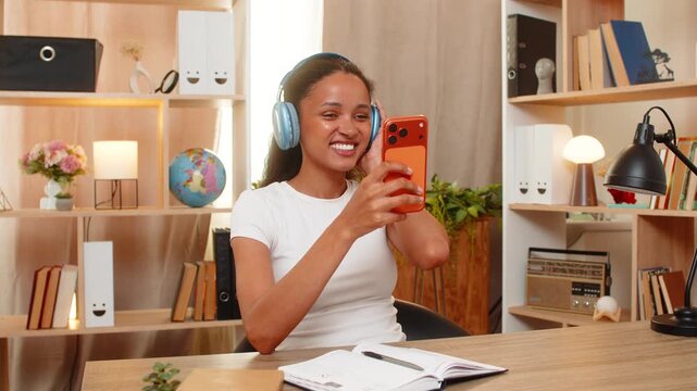 African American young woman on home table starts language lesson on smartphone and listens to phrase with headphones. Black girl learns new words and practices listening skills with notebook at home.